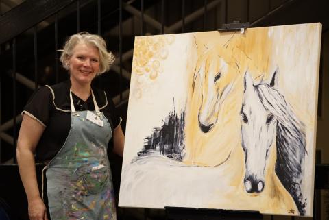 Person in art attired smiling with their recently completed painting of two horses
